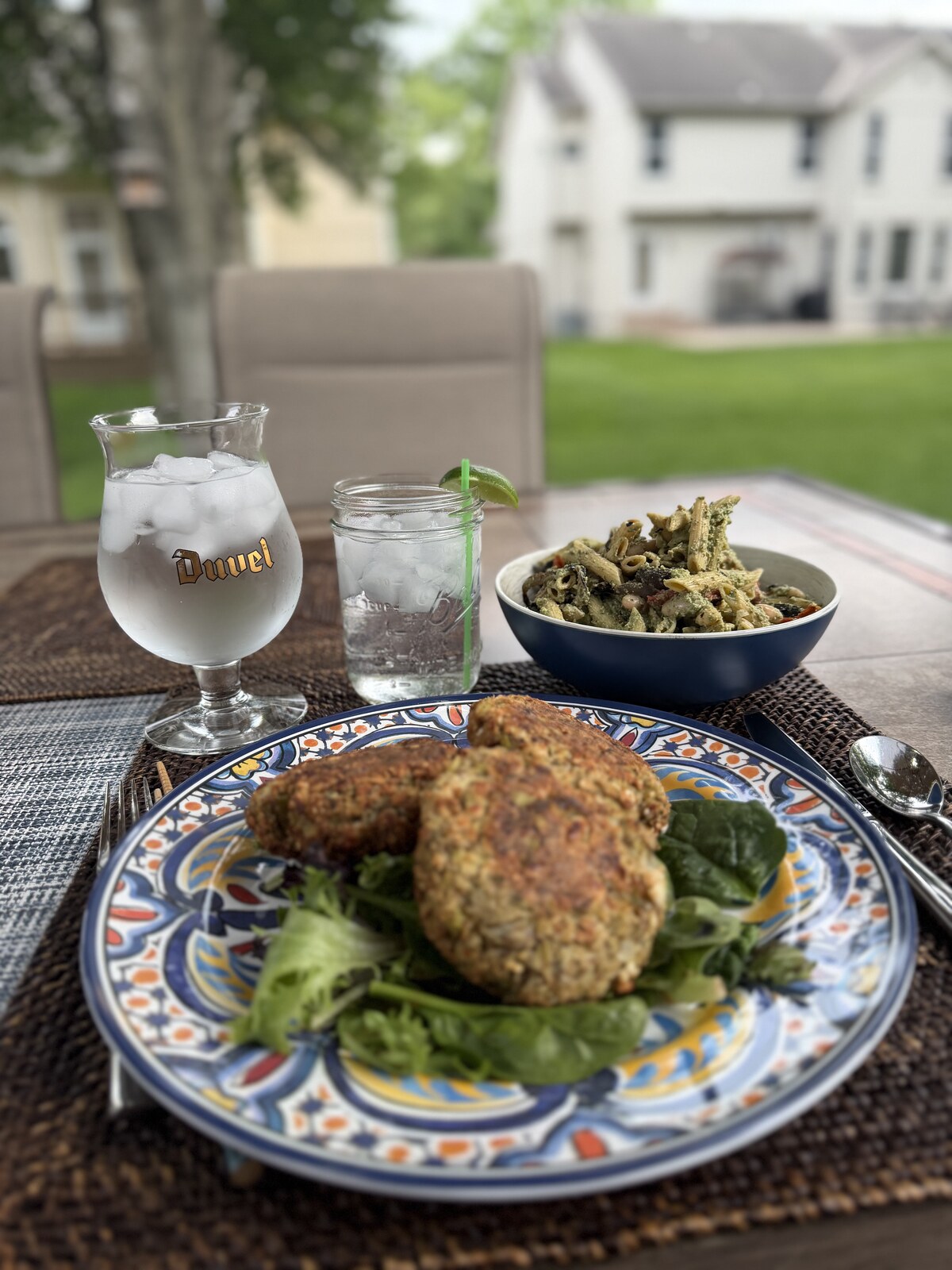Two golden-crusted white bean burger patties on a hand-painted plate with greens, served alongside a bowl of pesto penne on an outdoor table