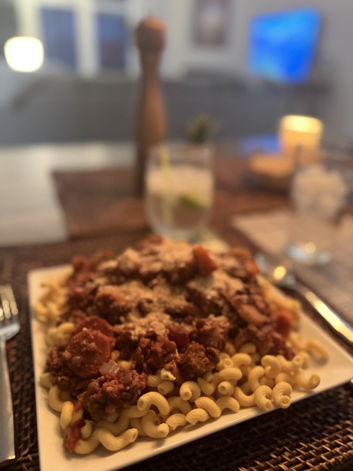 A white plate of cavatappi pasta topped with a hearty lentil and tomato bolognese, dusted with plant-based parmesan