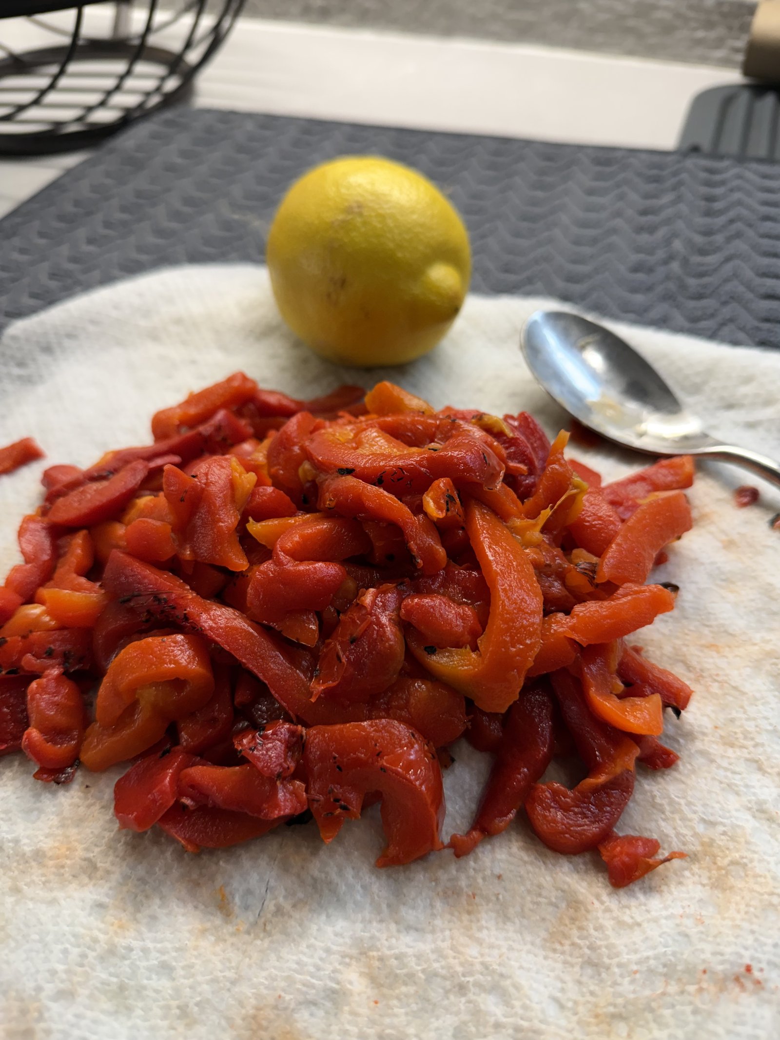 A pile of silky, freshly peeled roasted red pepper strips on a paper towel, with a whole lemon and a spoon beside them