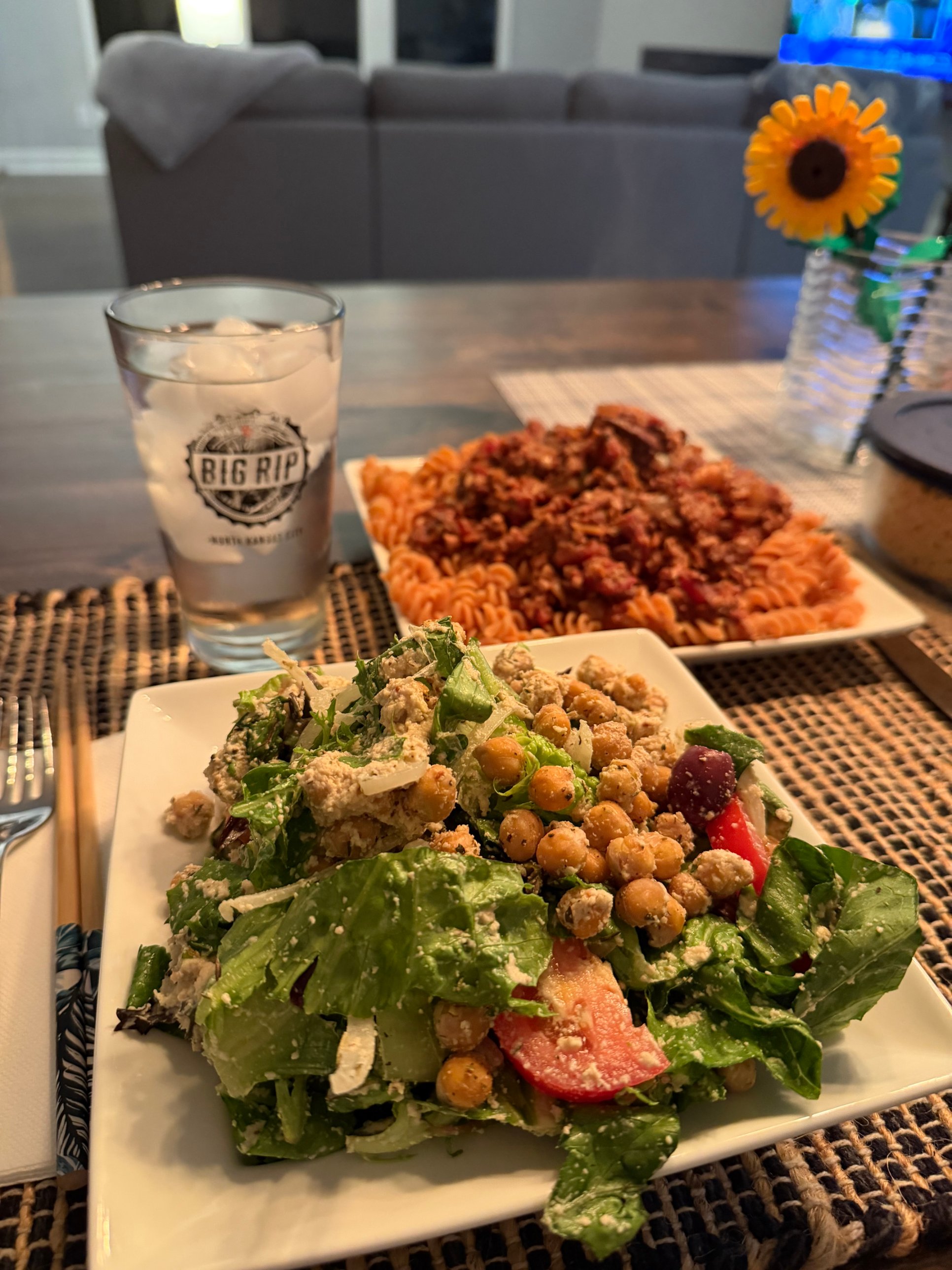 A square white plate of chopped romaine salad with crispy herb-roasted chickpeas, tomato wedges, Kalamata olives, shaved onion, and a creamy cashew dressing in the foreground, with a plate of red lentil rotini topped with a hearty vegan bolognese waiting behind it next to a glass of ice water