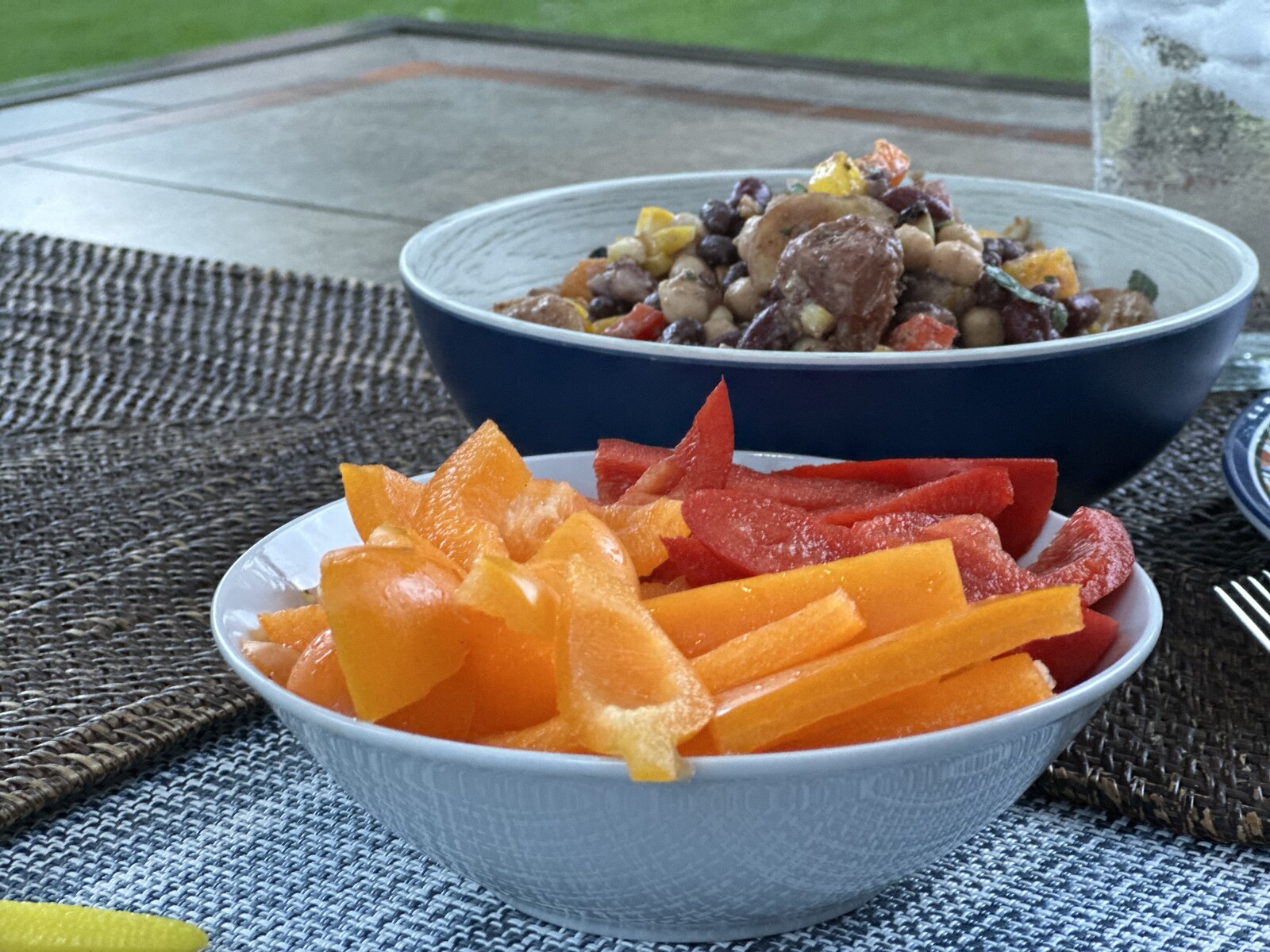 A navy bowl of three-bean and baby potato salad with sweet corn and fresh herbs, served with a side bowl of sliced red and orange bell peppers on a patio table