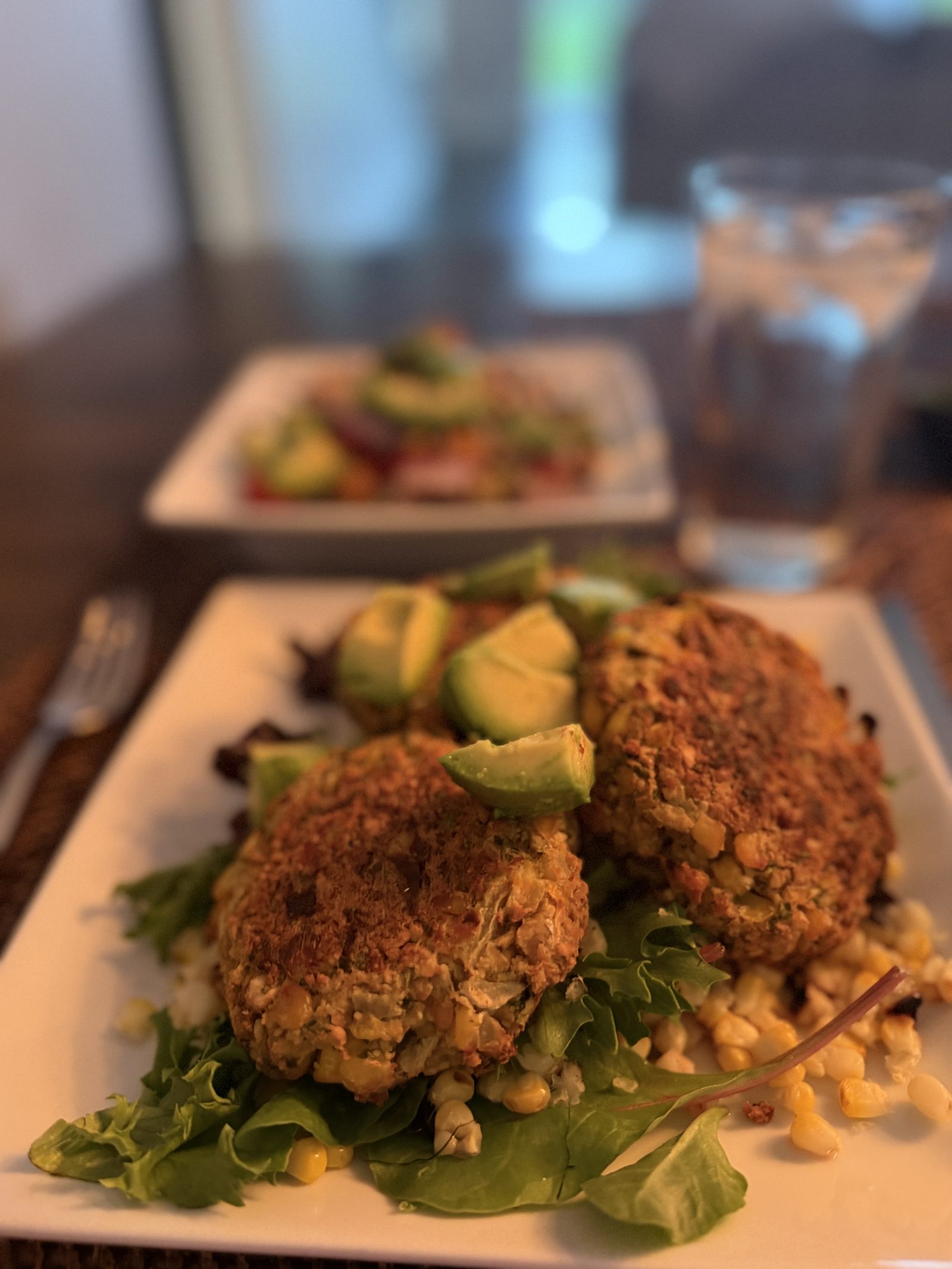 Two golden pan-seared corn cakes over baby greens with grilled corn kernels and avocado wedges, a bowl of black bean rice salad blurred in the background
