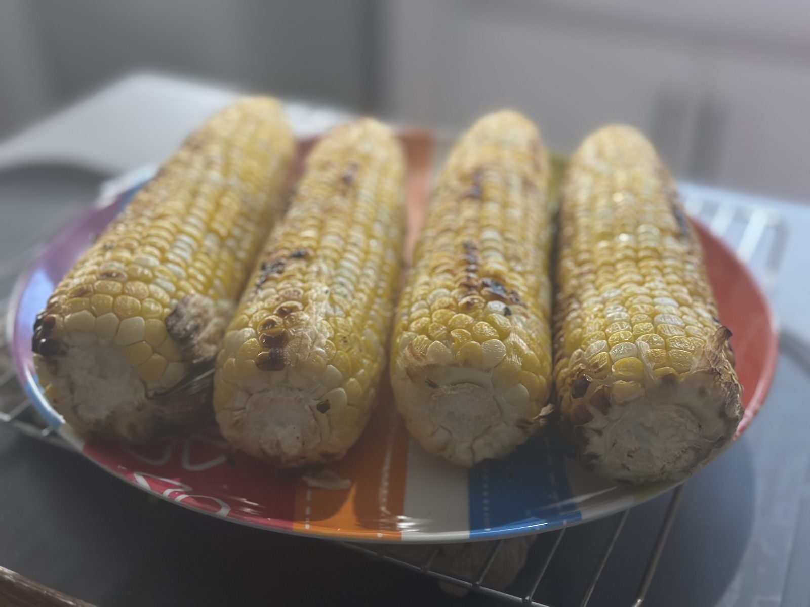Four ears of grilled sweet corn, charred and glistening, piled on a colorful striped plate