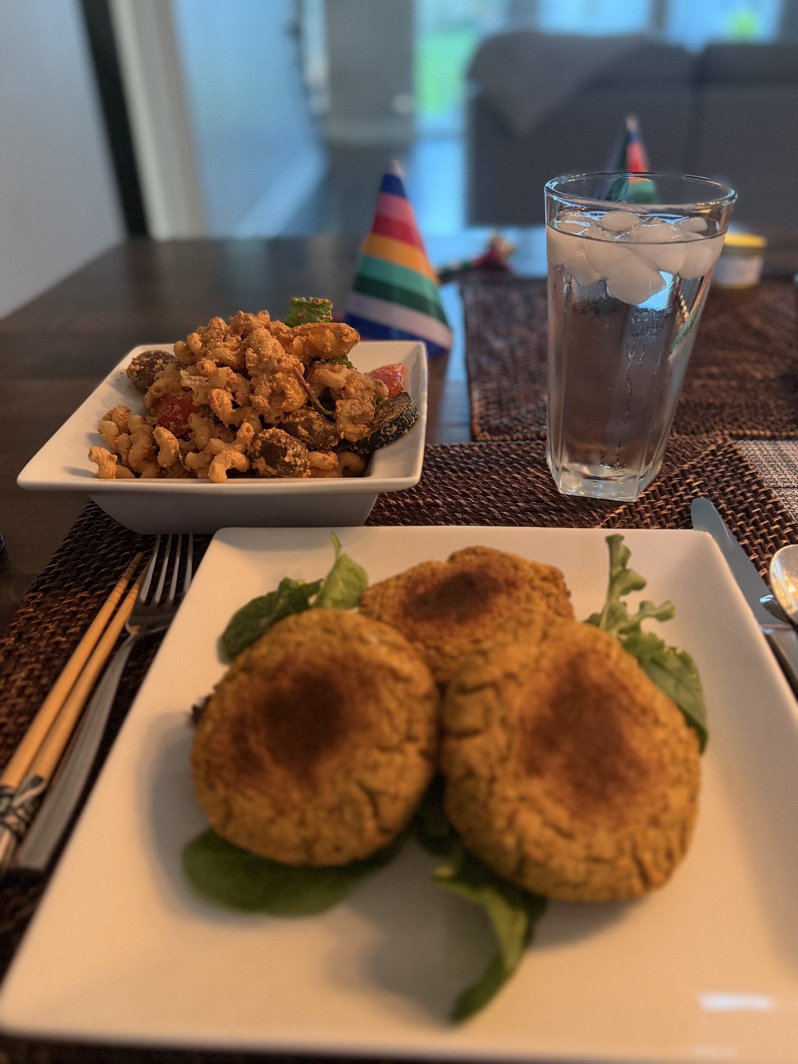 Three golden pan-seared red pepper falafel patties on a bed of mixed greens, served alongside a bowl of macaroni pasta salad with Kalamata olive hummus, cherry tomatoes, olives, and herbs