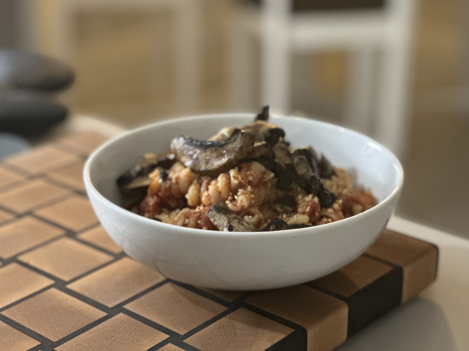 A white bowl of twisted pasta in a rustic tomato sauce, piled with roasted portobello slices, white beans, and a dusting of golden plant-based parmesan