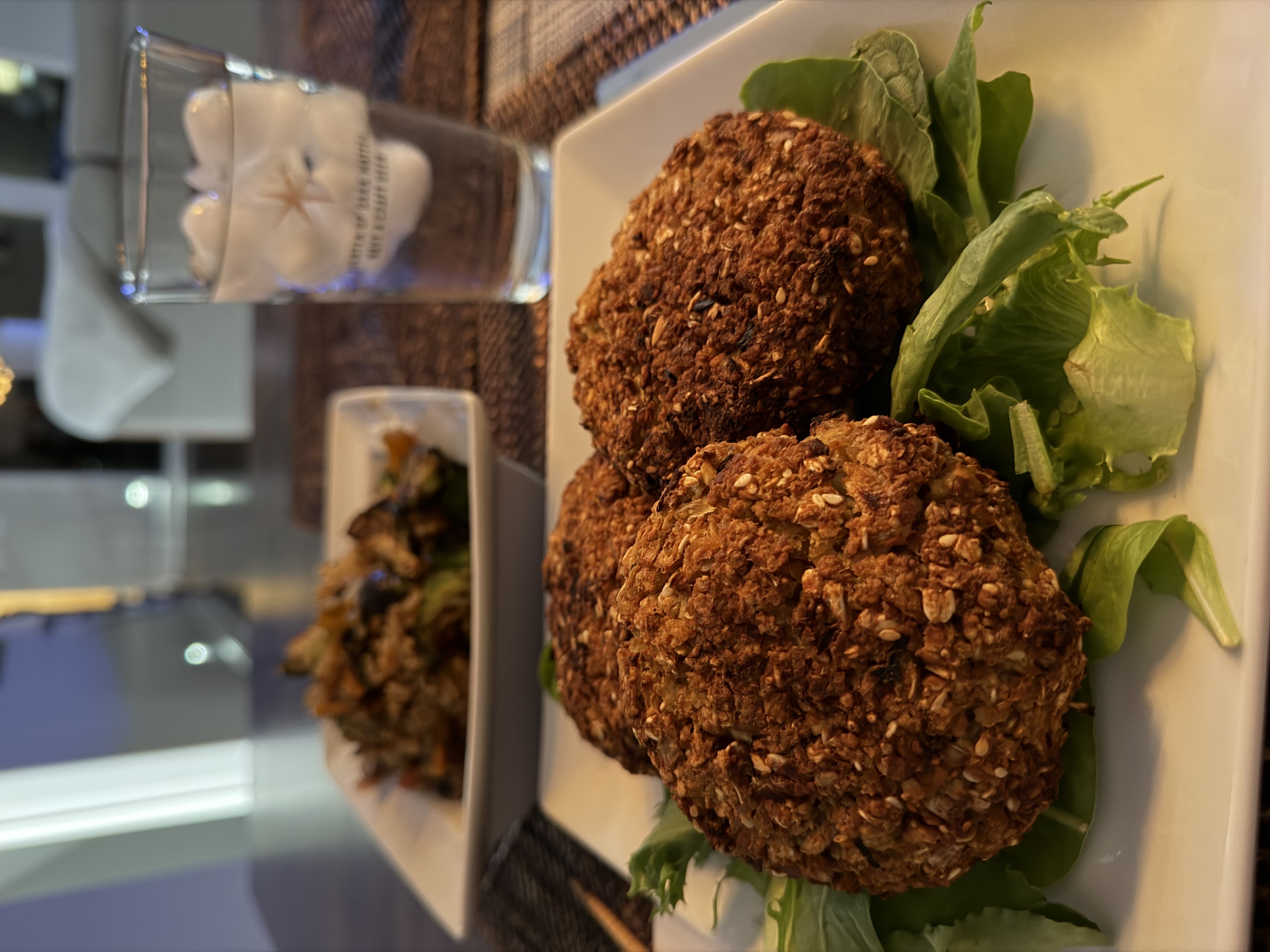 Three golden, oat-and-sesame-crusted teriyaki burger patties over crisp lettuce, with a bowl of teriyaki fried rice and charred brussels sprouts blurred in the background, alongside a bottle of teriyaki sauce and a glass of ice water